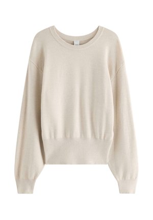 Ung kvinde med langt blondt hår iført en beige sweater, mørkeblå vide jeans og beige sneakers, stående mod en ensfarvet baggrund.