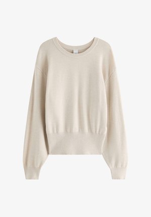 Ung kvinde med langt blondt hår iført en beige sweater, mørkeblå vide jeans og beige sneakers, stående mod en ensfarvet baggrund.