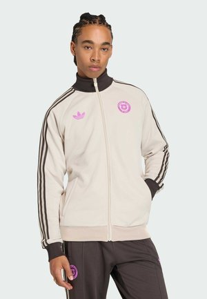 Personne portant une veste et un pantalon de sport beige et marron foncé avec des logos roses et des bandes noires sur les manches et les côtés du pantalon.