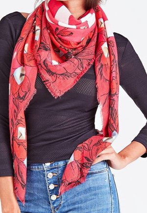 Femme portant une écharpe rouge à fleurs avec des bords frangés sur un haut noir texturé et un jean bleu taille haute avec des boutons blancs.