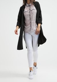 Černý dlouhý cardigan přes vzorovanou blůzu s límcem, doplněný o bílé skinny džíny a bílé tenisky. Hladká tkanina, volný střih.