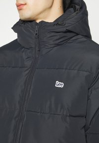 Doudoune noire avec un col montant, fermeture éclair et capuche ajustable. Présente un petit logo "Lee" blanc sur la poitrine.