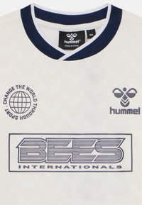 Witte sporttrui met marineblauwe accenten, voorzien van een chevronpatroon, een rond logo met de tekst "Verander de Wereld door Sport" en de tekst "BEES INTERNATIONALS".