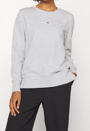 Kvinde iført en lysegrå Tommy Hilfiger sweatshirt med logo på brystet og sorte bukser, stående med den ene hånd i lommen.