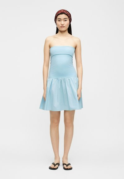 Νεαρή γυναίκα που φοράει αμάνικο φωτεινό γαλάζιο φόρεμα, κόκκινη κορδέλα με σχέδια στα μαλλιά και μαύρα σαγιονάρες, στέκεται μπροστά σε ένα απλό λευκό φόντο.