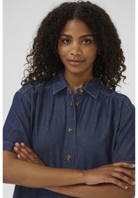 Mørkeblå denimskjorte med korte ærmer, knaplukning og skr gathered detalje ved skuldrene, kombineret med naturligt, krøllet hår.