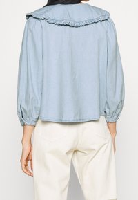 Chemise en denim bleu clair avec un col à volants, des manches longues avec un ourlet à poignets et une coupe décontractée. Le tissu semble doux avec des détails de couture visibles.