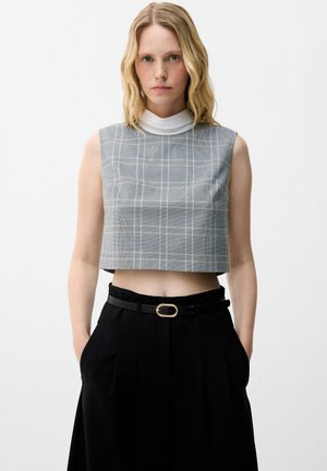 Jeune femme portant un crop top à carreaux gris sans manches sur un col roulé blanc et un pantalon noir taille haute avec ceinture, debout devant un fond blanc.