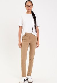 Pantalons en coton beige avec une coupe ajustée, dotés d'une fermeture à bouton et de poches, associés à un t-shirt blanc uni et des baskets.