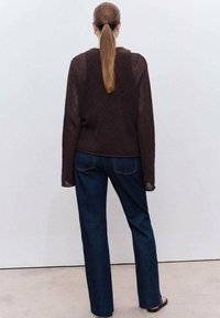 Mujer con una coleta larga que lleva un suéter de malla marrón oscuro y unos vaqueros azul oscuro, de pie contra una pared gris clara y lisa.