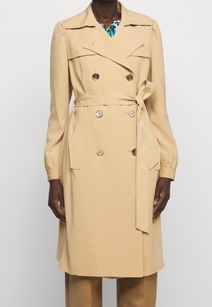 Persona con un abrigo beige de doble botonadura con cinturón en la cintura y una camisa con estampado floral visible en el cuello.