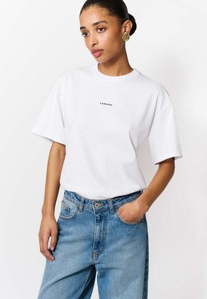 Kobieta z zaczesanymi do tyłu włosami, nosząca złote kolczyki, białą koszulkę LAMANIA oraz niebieskie dżinsy, z jedną ręką w kieszeni, na białym tle.