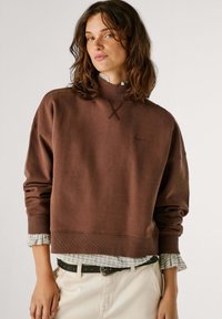 Hnedá oversized mikina s vysokým golierom a rebrovanými manžetami, vrstvená nad kockovanou košeľou. Obsahuje decentné logo vyšívané vpredu.