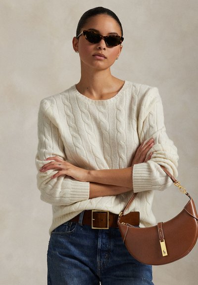 Kvinde iført skildpaddemønstrede solbriller, cremefarvet kabelstrikket sweater, blå jeans, brun bælte og holder en brun halvmåneformet håndtaske.