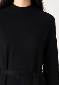 Musta pitkähihaine neule, jossa on ribbed kaulus, pehmeä tekstuuri, istuva muotoilu ja vyöllä varustettu yksityiskohta. Minimalistinen ja elegantti ilme.