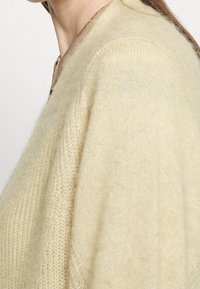 Beige stickad cardigan gjord av ett mjukt, texturerat material. Har en rund halsringning och synlig sömnad längs kanterna.