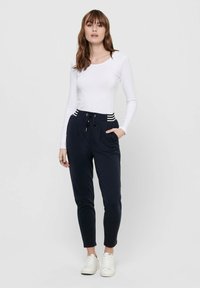 Weißes Langarmoberteil kombiniert mit marineblauen Jogginghosen mit Kordelzug und gestreiften Akzenten. Das Modell steht in weißen Sneakern.