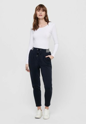 White long-sleeve top paired with navy jogger pants featuring a drawstring waist and striped accents. Model stands in white sneakers.