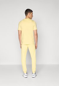 Chemise jaune à manches courtes et pantalon de jogging assorti, avec un design ajusté, une texture douce et des poignets côtelés, porté avec des chaussures de sport blanches.