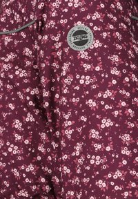 Blommönstrad tyg i vinröd med rosa och vita blommor. Har en rund logotyp med texten "ZIGZAG" och grå trimaccent.