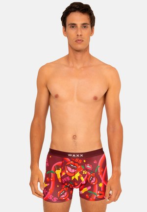 Jeune homme portant un boxer coloré avec des motifs de lèvres, cerises, fraises, rouge à lèvres et cœurs sur un fond clair uni.