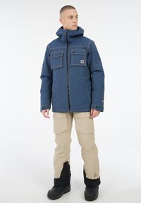 Mann in blauer Kapuzenjacke und beigen Cargo-Shorts mit schwarzen Schneestiefeln, der vor einem weißen Hintergrund steht und nach rechts schaut.