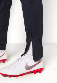 Marina atletiska byxor med dragkedjeöppningar vid anklarna, i kombination med vita och röda Nike fotbollsskor med en silverfärgad swoosh och texturerad ovandel.