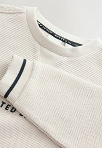 Sudadera de un beige claro con un patrón de tejido texturizado, que presenta un cuello redondo acanalado y acentos en negro en el puño. Branding visible.