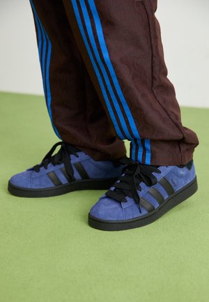 Siniset mokkakengät, joissa on mustat raidat, musta pohja ja mustat nauhat. Ruskeat kuvioidut housut, joissa on siniset sivuraidat. Näkyvissä vihreä lattiatekstuuri.