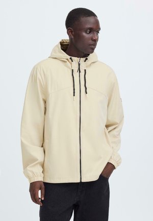 Mand iført en beige lynlåstrøje med hætte og sorte snore, poserende med den ene hånd i lommen og kigger til siden.