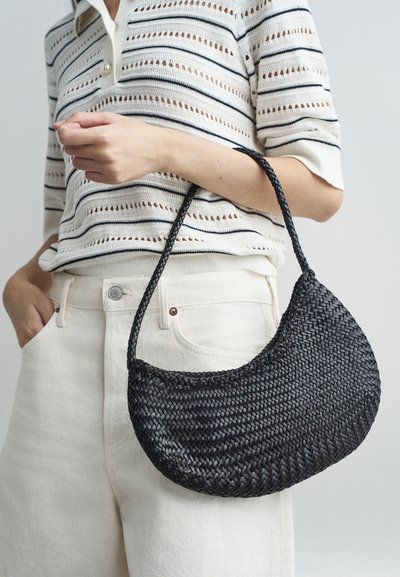 Μαύρη πλεκτή τσάντα σε σχήμα ημισελήνου με πλεχτό χερούλι, εκτεθειμένη σε ανοιχτό φόντο, συνδυασμένη με καρό πλεκτό μπλουζάκι και λευκά παντελόνια.