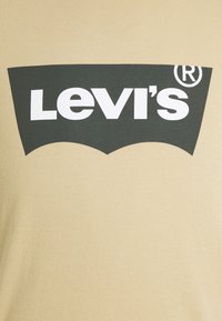 Logo Levi’s en blanc sur une forme trapézoïdale foncée imprimée sur un tissu beige.