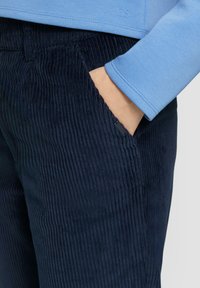Navy corduroy broeken met verticale ribbels, voorzien van een zijzak. Een lichtblauw langemouwen shirt met een gladde textuur is gedeeltelijk zichtbaar.