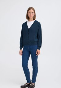 Cardigan lavorato a maglia blu navy con scollo a V, maniche lunghe e chiusura con bottoni, abbinato a jeans skinny blu e scarpe scure. Texture liscia.