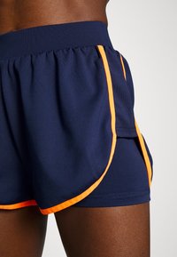 Mitad inferior de una persona que lleva pantalones cortos deportivos azul marino con un brillante ribete naranja, sobre un fondo claro y liso.