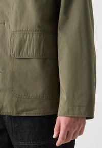 Olive green, lightweight jacket with a textured finish, featuring a side pocket and a straight-cut sleeve. Black denim visible at the bottom.