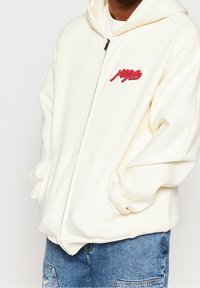 Witte hoodie met ritssluiting, gemaakt van zacht materiaal, voorzien van een rood geborduurd logo op de borst. Kangeroezak en verstelbare trekkoord capuchon.