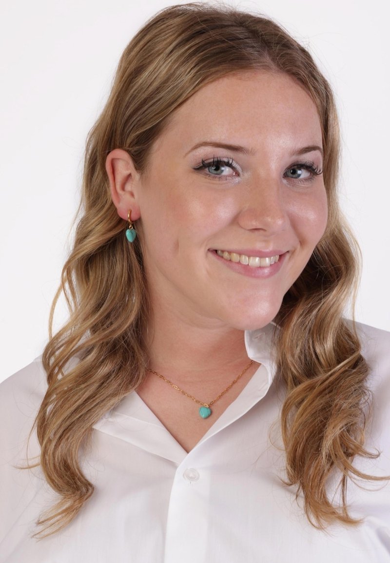 Gouden ketting met een turquoise hanger, gecombineerd met bijpassende turquoise oorknoppen. Model draagt een witte button-up blouse.
