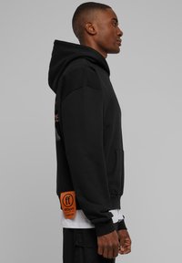 Fekete kapucnis pulóver kenguru zsebbel, a szegélyen fehér szöveggel ellátott narancssárga címkével. Simaságú anyag, kényelmes szabás és klasszikus kialakítás.