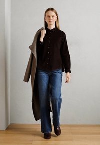 Woman walking indoors wearing a black button-up shirt, blue jeans, brown shoes, and carrying a long brown coat over her shoulder.