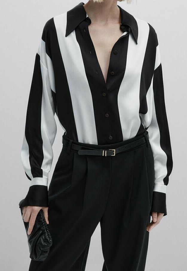 Personne portant une chemise à rayures verticales noires et blanches rentrée dans un pantalon noir taille haute avec une ceinture noire, tenant une pochette noire.