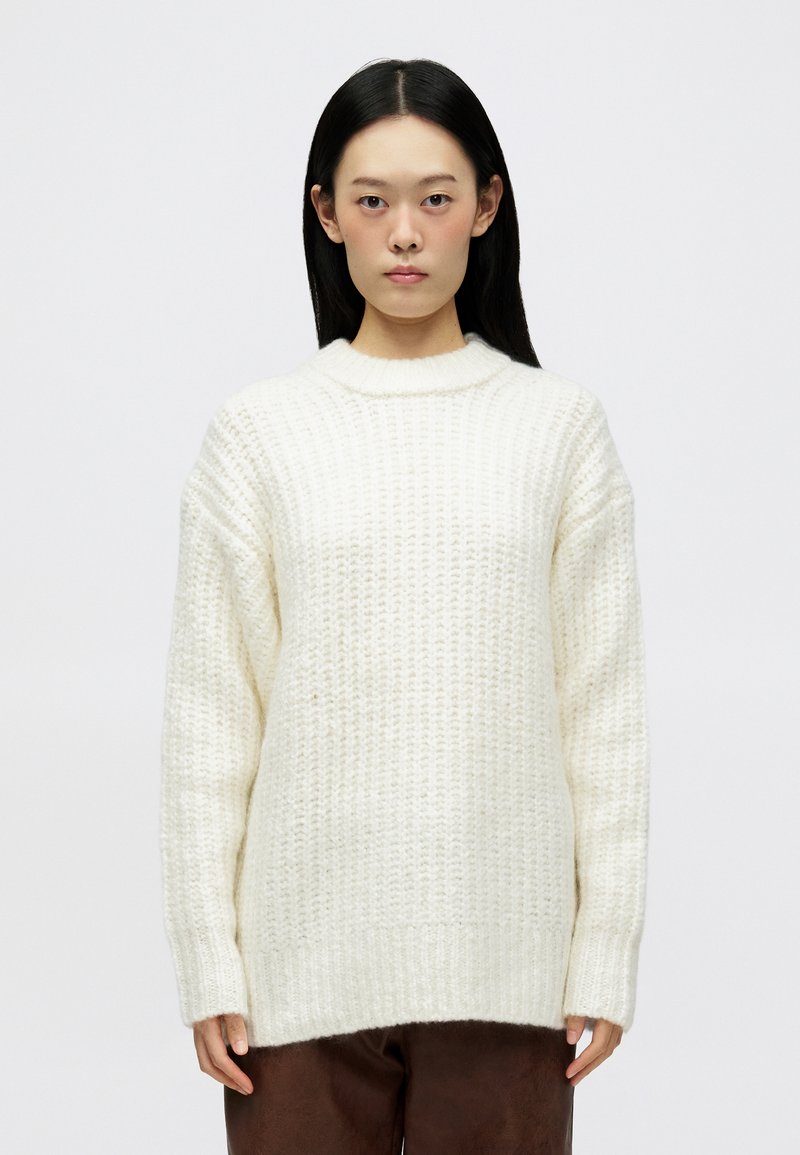 Λευκό πλεκτό πουλόβερ με χοντρή υφή, χαλαρή γραμμή και ριμπ κοψίματος στον λαιμό. Μακριά μανίκια και ελαφρώς υπερβολικό σχήμα.