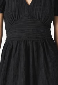 Μαύρο φόρεμα με υφή από ζαρωμένο ύφασμα. V-λαιμόκοψη και πτυχώσεις στη μέση, ρέουσα σε φαρδύ φούστα.