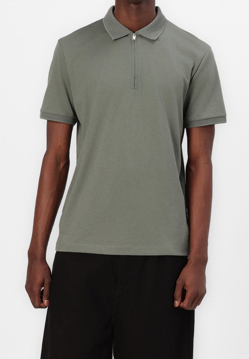 Polo à manches courtes vert olive en tissu doux. Il présente une fermeture à demi-zip à l'avant et un col texturé. Design simple sans motifs.