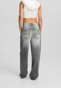 Nainen, joka pitää yllä väljät haalistuneet harmaat farkut ja valkoista hihattomaa cropped-hupparia, seisoo käsivarret takataskuissa yksinkertaista taustaa vasten.