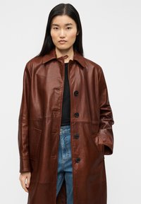 Καφέ δερμάτινο παλτό με γιακά, κουμπιά μπροστά και πλαϊνές τσέπες, με κομψή υφή και ίσια, μακριά σχεδίαση.