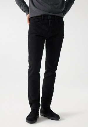 Schwarze Jeans mit Slim-Fit, ausgestattet mit einem klassischen Fünf-Taschen-Design und einer dezenten Textur. Kombiniert mit schwarzen Wildleder-Stiefeletten.