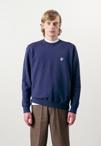 Sweatshirt in cotone navy con polsini e orlo a coste, caratterizzato da un piccolo logo sul petto, abbinato a pantaloni larghi di colore marrone chiaro.