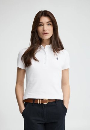 Kobieta o długich brązowych włosach, ubrana w biały polo z krótkim rękawem, brązowy pasek i czarne spodnie, stojąca z rękami w kieszeniach.