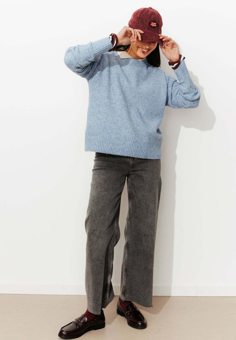 Pull en tricot bleu clair avec un ourlet côtelé, associé à un jean gris à taille large avec des zips sur les côtés, et des mocassins sombres. Casquette bordeaux incluse.